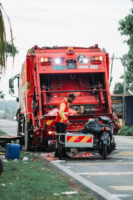 A waste collection worker wearing a high-visibility red and yellow uniform is standing on a paved roadside pavement, operating the rear loading mechanism of a large red garbage truck. The truck is par